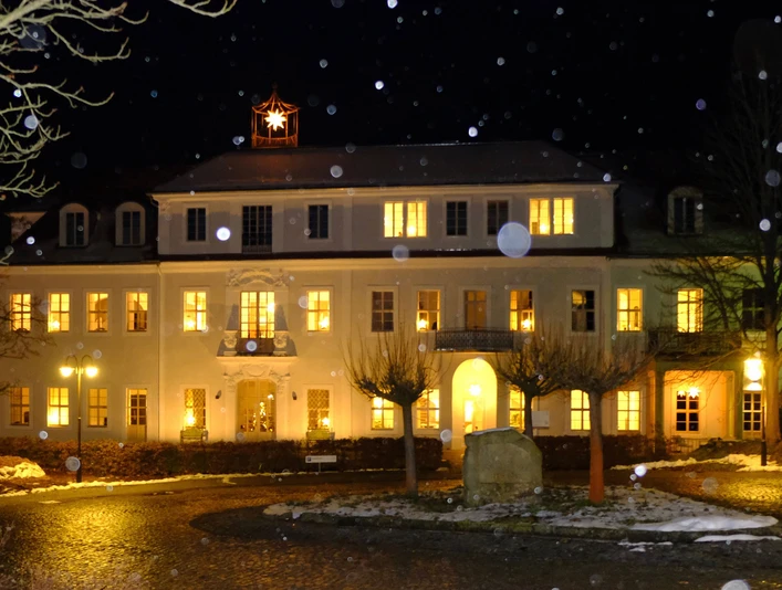 Ein beleuchtetes, zweistöckiges Gebäude bei Nacht mit einem leuchtenden Stern auf dem Dach; Schneeflocken fallen, warme Atmosphäre.An illuminated, two-story building at night with a glowing star on the roof; snowflakes falling, warm atmosphere.Osvětlená dvoupatrová budova v noci se zářící hvězdou na střeše; padající sněhové vločky, hřejivá atmosféra.Oświetlony, dwupiętrowy budynek nocą ze świecącą gwiazdą na dachu; spadające płatki śniegu, ciepła atmosfera.Een verlicht gebouw van twee verdiepingen 's nachts met een stralende ster op het dak; vallende sneeuwvlokken, warme sfeer.Un edificio a due piani illuminato di notte con una stella splendente sul tetto; fiocchi di neve che cadono, atmosfera calda.
