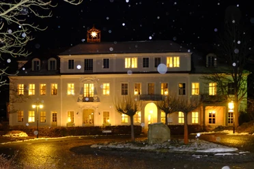 Schloss-Prossen Ein beleuchtetes, zweistöckiges Gebäude bei Nacht mit einem leuchtenden Stern auf dem Dach; Schneeflocken fallen, warme Atmosphäre.
