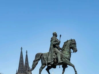 Reiterstandbild König Friedrich Wilhelm III. Das Bild zeigt das bronzene Reiterstandbild von König Friedrich Wilhelm III. auf einem steinernen Sockel. Der detailreich gearbeitete König sitzt mit stolzer Haltung auf einem majestätischen Pferd. Entlang des Sockels sind ebenfalls bronzene Figuren abgebildet, die bedeutende Persönlichkeiten jener Epoche darstellen. Im Hintergrund sind die beiden markanten Türme des Kölner Doms erkennbar. The picture shows the bronze equestrian statue of King Frederick William III on a stone pedestal. The richly detailed king sits proudly on a majestic horse. Along the pedestal are also bronze figures depicting important personalities of the era. The two striking towers of Cologne Cathedral can be seen in the background.