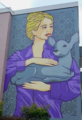 Mural A mural shows a woman with blonde hair holding a deer in a purple jacket. This is a work by the artist El Bocho, which can be admired on the façade of a house in Cologne.