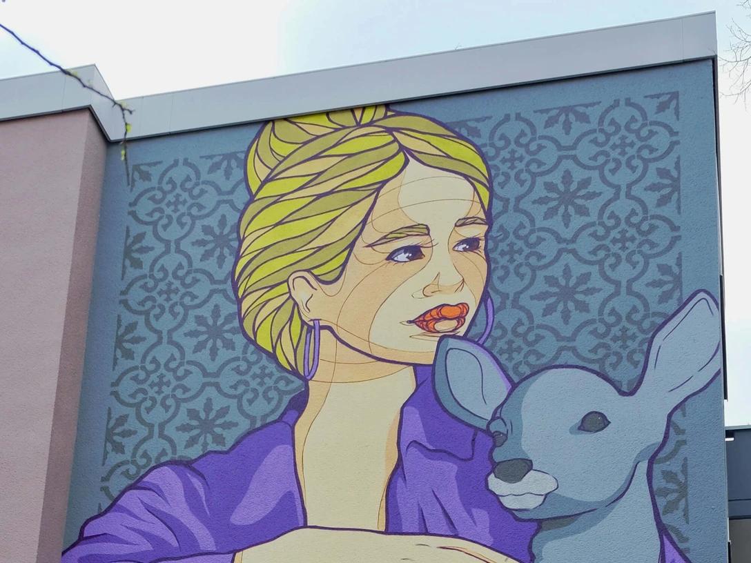 Mural Eine Wandmalerei zeigt eine Frau mit blonden Haaren, die in einer lila Jacke ein Reh hält. Dies ist ein Werk des Künstlers El Bocho, das an einer Hausfassade in Köln zu bewundern ist.A mural shows a woman with blonde hair holding a deer in a purple jacket. This is a work by the artist El Bocho, which can be admired on the façade of a house in Cologne.