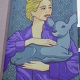 Mural Eine Wandmalerei zeigt eine Frau mit blonden Haaren, die in einer lila Jacke ein Reh hält. Dies ist ein Werk des Künstlers El Bocho, das an einer Hausfassade in Köln zu bewundern ist.A mural shows a woman with blonde hair holding a deer in a purple jacket. This is a work by the artist El Bocho, which can be admired on the façade of a house in Cologne.