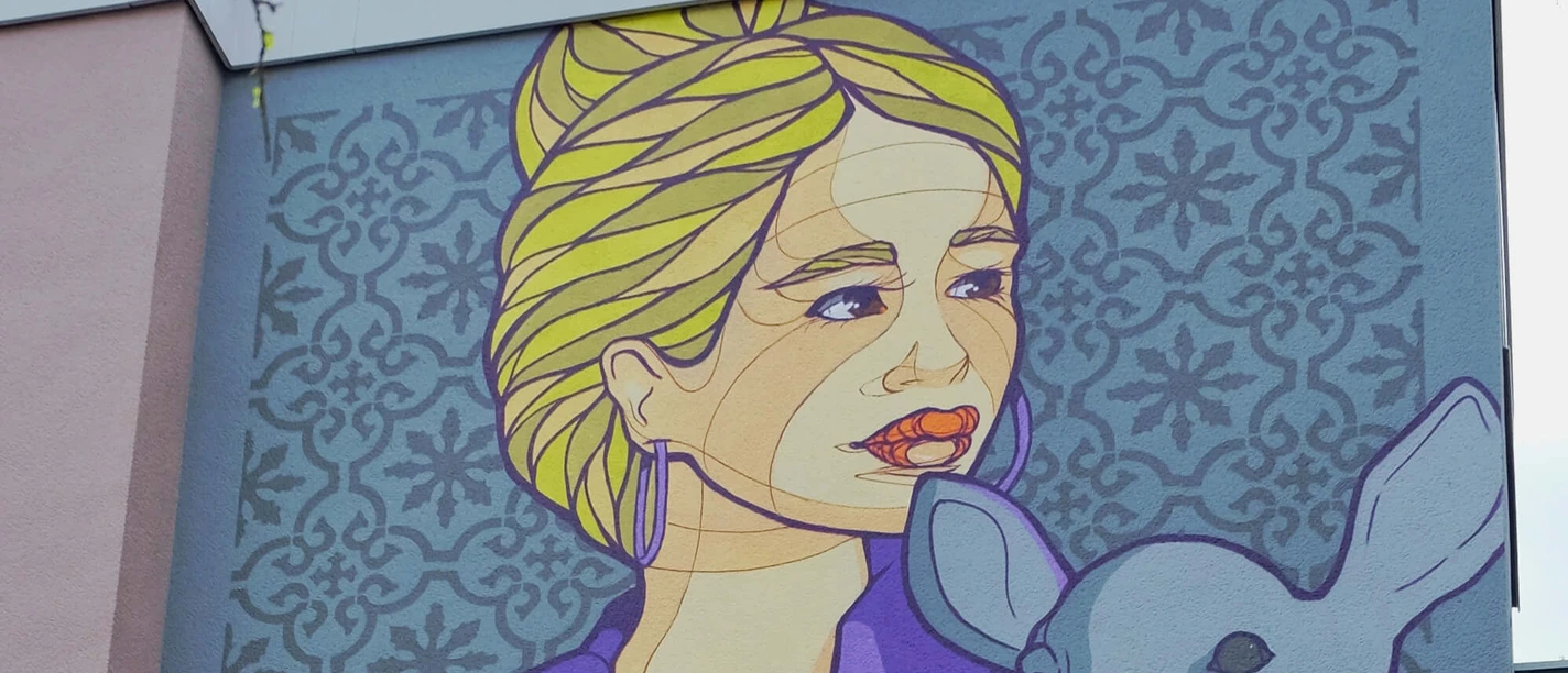 Mural A mural shows a woman with blonde hair holding a deer in a purple jacket. This is a work by the artist El Bocho, which can be admired on the façade of a house in Cologne.