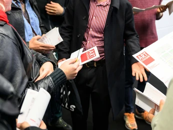 ART COLOGNE Gruppe von Menschen hält Flugblätter mit "ART COLOGNE"-Logo und diskutiert angeregt.Group of people holding leaflets with the "ART COLOGNE" logo and having a lively discussion.