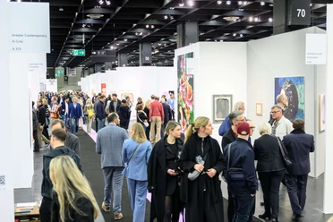 ART COLOGNE Menschen flanieren entlang der Stände, betrachten aufmerksam verschiedene Kunstwerke, die an weißen Wänden präsentiert werden. Im Vordergrund zieht eine bunte Mischung aus Gemälden die Blicke der Betrachter*innen auf sich, während die Decke durch industrielle Beleuchtung hervorsticht. Die Szene wirkt lebendig und zeigt das Interesse der Menschen an zeitgenössischer Kunst.