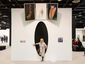 ART COLOGNE Ein geräumiger weißer Ausstellungsraum mit ovaler Öffnung, aus der eine Person hervorkommt; darüber sind drei Gemälde zu sehen. A spacious white exhibition room with an oval opening from which a person emerges; three paintings can be seen above.