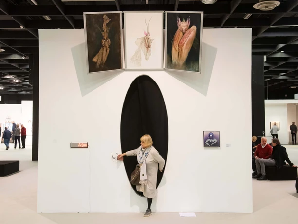 ART COLOGNE A spacious white exhibition room with an oval opening from which a person emerges; three paintings can be seen above.