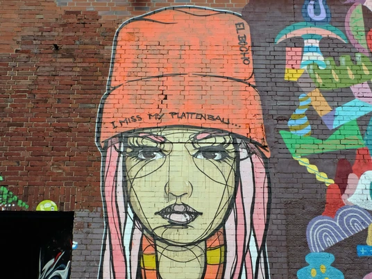 Mural Das Graffiti zeigt ein großflächiges Bild einer Frau mit pinken Haaren, einer orangefarbenen Mütze mit der Aufschrift "I miss my Plattenbau" und einem gelb-orange gestreiften Shirt. Der Hintergrund ist eine rote Backsteinwand, daneben farbenfrohe, abstrakte Elemente.The graffiti shows a large image of a woman with pink hair, an orange cap with the words "I miss my Plattenbau" and a yellow and orange striped shirt. The background is a red brick wall with colorful, abstract elements next to it.