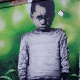 Mural Ein großformatiges Graffiti zeigt einen jungen Jungen mit ernstem Blick vor einem grünen Hintergrund. Auf seinem Pullover steht die verblasste Botschaft: „I WILL NEVER SAY WHAT YOU WANT TO HEAR“.A large-format graffiti shows a young boy with a serious look in front of a green background. The faded message on his sweater reads: "I WILL NEVER SAY WHAT YOU WANT TO HEAR".