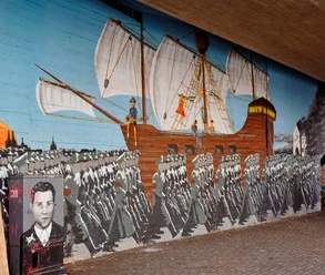 Mural Ein großflächiges Wandgemälde zeigt eine historische Szene der Edelweißpiraten in Köln. Ein Segelschiff mit weißen Segeln zieht über den Ozean, während im Vordergrund eine marschierende Menschenmenge zu sehen ist. Am linken Bildrand ist ein Porträt eines jungen Mannes zu erkennen. Das lebendige, historische Kunstwerk ziert eine Unterführung und erinnert an die soziale Geschichte der Stadt.