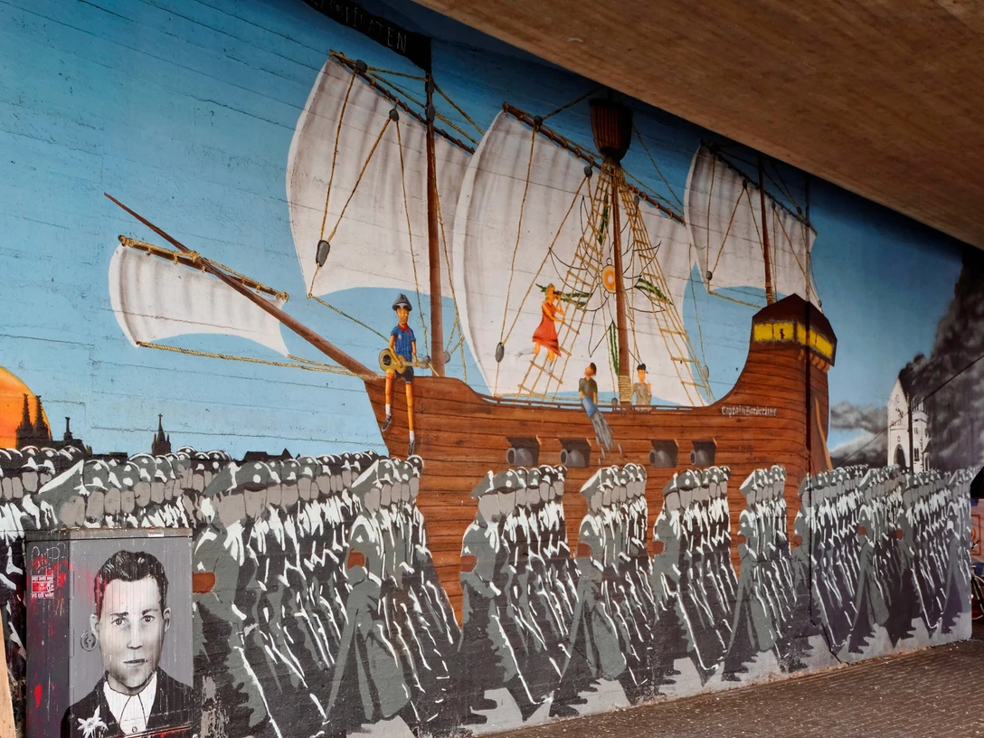 Mural Ein großflächiges Wandgemälde zeigt eine historische Szene der Edelweißpiraten in Köln. Ein Segelschiff mit weißen Segeln zieht über den Ozean, während im Vordergrund eine marschierende Menschenmenge zu sehen ist. Am linken Bildrand ist ein Porträt eines jungen Mannes zu erkennen. Das lebendige, historische Kunstwerk ziert eine Unterführung und erinnert an die soziale Geschichte der Stadt.A large-scale mural shows a historical scene of the Edelweiss Pirates in Cologne. A sailing ship with white sails is sailing across the ocean, while a marching crowd can be seen in the foreground. A portrait of a young man can be seen on the left-hand edge of the picture. The lively, historical work of art adorns an underpass and is a reminder of the city's social history.