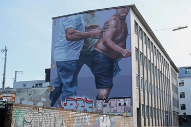 Mural Das Bild zeigt ein großflächiges Wandgemälde auf einem mehrstöckigen Gebäude. Zwei männliche Figuren sind in einer dynamischen Bewegung dargestellt, wobei eine Person ein blaues T-Shirt trägt und die andere, halbnackte, von dieser gestützt wird.