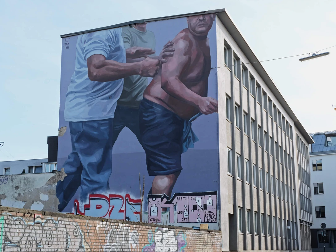 Mural Das Bild zeigt ein großflächiges Wandgemälde auf einem mehrstöckigen Gebäude. Zwei männliche Figuren sind in einer dynamischen Bewegung dargestellt, wobei eine Person ein blaues T-Shirt trägt und die andere, halbnackte, von dieser gestützt wird.The picture shows a large-scale mural on a multi-storey building. Two male figures are depicted in a dynamic movement, with one person wearing a blue T-shirt and the other, half-naked, being supported by it.