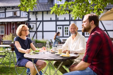 Biergarten Rusticus Leute sitzen entspannt an einem Tisch im Freien, genießen Essen und Trinken unter Bäumen.