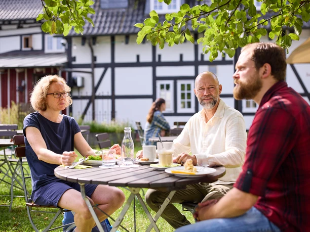 Biergarten Rusticus Leute sitzen entspannt an einem Tisch im Freien, genießen Essen und Trinken unter Bäumen.