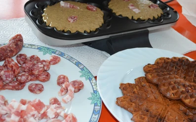 Speckendicken zu Silvester Geopend wafelijzer met beslag en stukjes spek, daarnaast een bord met salami, spek en afgewerkte wafels.