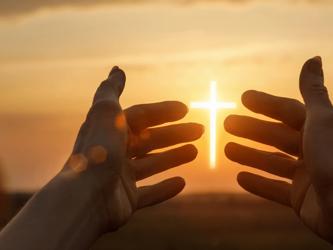 Bethaus Das Bild zeigt einen Sonnenuntergang im Hintergrund. Zwei Hände werden hochgehalten, und zwischen ihnen ist das Jesuskreuz zu sehen, das die Sonne darstellt.