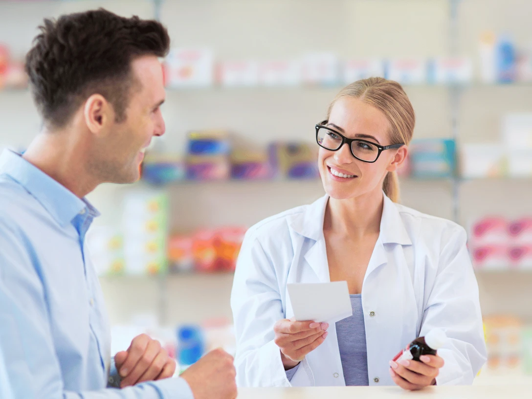 Brunnenapotheke Destination one.png Das Bild zeigt eine Apothekerin, die einen Kunden freundlich berät und dabei lächelt. Im Hintergrund sind Regale mit weiteren Gesundheitsprodukten und Medikamenten zu sehen.