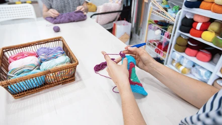 Diddi´s Hobbytreff.png Person strickt farbenfrohes Werkstück, während bunte Wollknäuel zur Auswahl stehen.