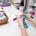 Diddi´s Hobbytreff.png Person strickt farbenfrohes Werkstück, während bunte Wollknäuel zur Auswahl stehen.