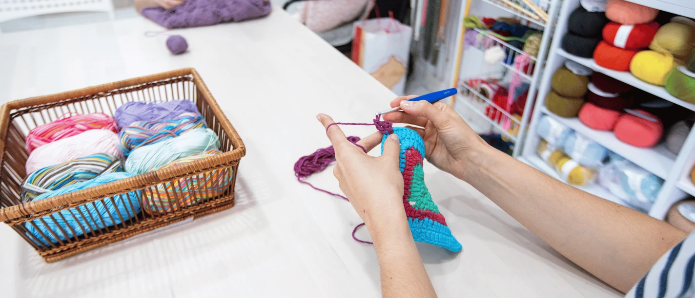 Diddi´s Hobbytreff.png Person strickt farbenfrohes Werkstück, während bunte Wollknäuel zur Auswahl stehen.