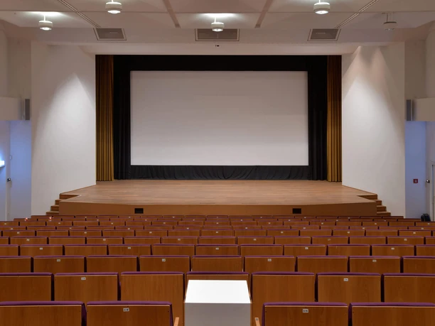 Bühne des Japanischen Kulturinstituts Holzvertäfelte Bühne im Japanischen Kulturinstitut mit Leinwand und roten Sitzplätzen.