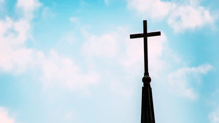 Evangelische Auferstehungskirche.png Eine Kirchturmspitze mit einem großen Kreuz ragt in den klaren, blauen Himmel.