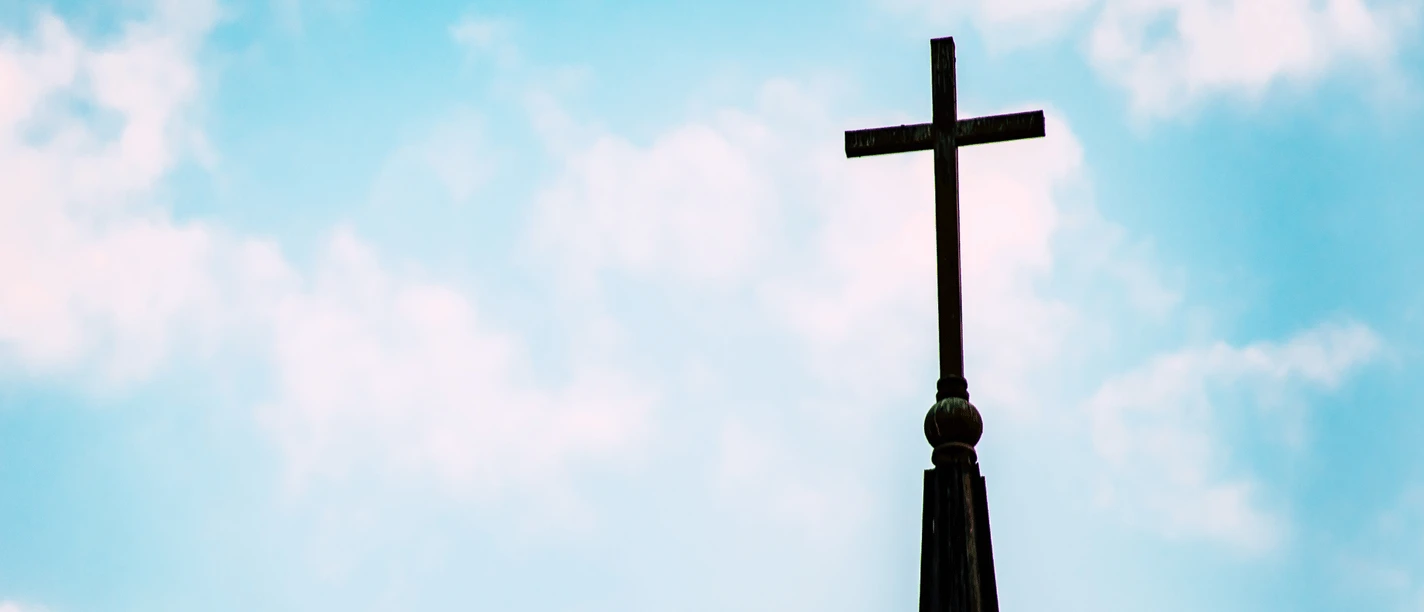 Evangelische Auferstehungskirche.png Eine Kirchturmspitze mit einem großen Kreuz ragt in den klaren, blauen Himmel.