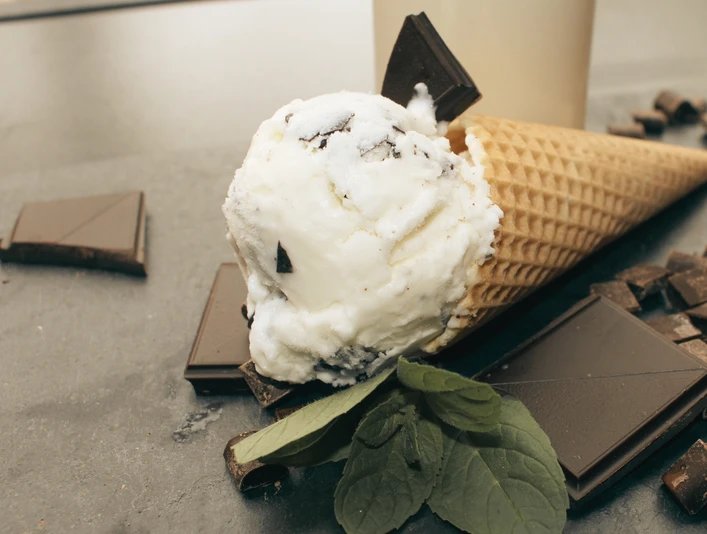 Gottschalk's Hof Stracciatellaeis Eine Eiswaffel mit Stracciatellaeis, umgeben von dunklen Schokoladenstücken und grünen Blättern.