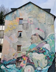 Mural Barrierefreie Beschreibung: Eine farbenfrohe Wandmalerei zeigt einen schlafenden Sufi mit Flügeln, umgeben von bunten Elementen.
