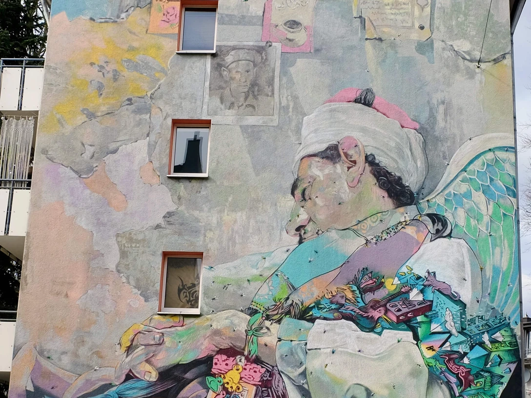 Mural Barrierefreie Beschreibung: Eine farbenfrohe Wandmalerei zeigt einen schlafenden Sufi mit Flügeln, umgeben von bunten Elementen.Accessible description: A colorful mural shows a sleeping Sufi with wings, surrounded by colorful elements.