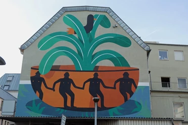 Mural Das Wandgemälde auf einer Hauswand zeigt vier figürlich stilisierte Personen in einer großen, pflanzenähnlichen Schale, umgeben von grünen und orangefarbenen Formen.