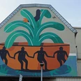 Mural Das Wandgemälde auf einer Hauswand zeigt vier figürlich stilisierte Personen in einer großen, pflanzenähnlichen Schale, umgeben von grünen und orangefarbenen Formen.The mural on a house wall shows four stylized figures in a large, plant-like bowl, surrounded by green and orange shapes.