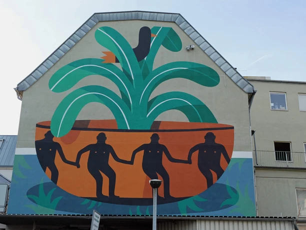 Mural Das Wandgemälde auf einer Hauswand zeigt vier figürlich stilisierte Personen in einer großen, pflanzenähnlichen Schale, umgeben von grünen und orangefarbenen Formen.