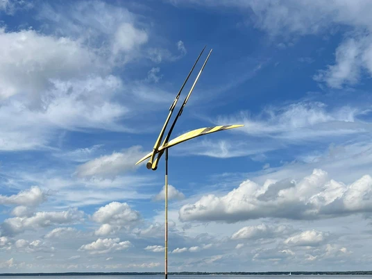 Skulptur vor der Promenade Libelle im Wind Skulptur mit filigranen Flügeln schwebt über ruhigem Wasser vor wolkigem Himmel.