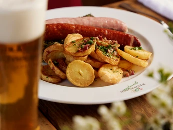 Bielefeld-gasthaus-buschkamp-bielefeldmarketing- silvioederling Ein Teller mit Bratkartoffeln, Bratwurst und Speck, begleitet von einem Glas Bier auf Holztisch.
