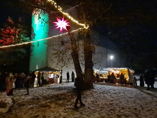 Löbnitzer Adventsmarkt Das Bild zeigt einen stimmungsvoll beleuchteten Weihnachtsmarkt bei Nacht, mit schneebedecktem Boden, geschmückten Verkaufsständen und einem markanten roten Stern, der in der Mitte zwischen den Bäumen hängt.