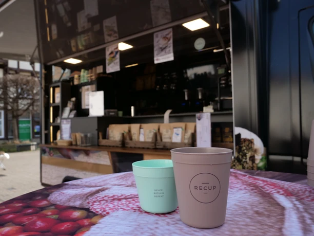 Ein brauner und ein grüner Pfandbecher der Marke Recup stehen vor dem Kaffeemobil auf einem Tisch