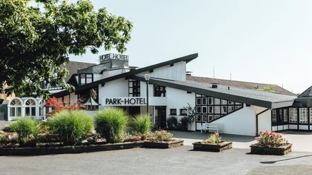 Haupteingang 2.jpg Modernes Parkhotel mit schrägen Dächern, umgeben von gepflegten Beeten und Pflanzen im Vordergrund.