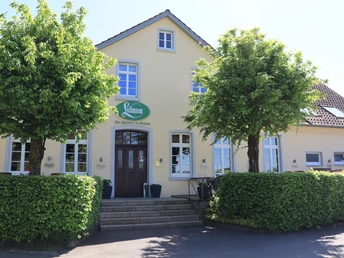 Zweistöckiges Gebäude mit gelber Fassade, brauner Tür, grünen Schild und flankierenden Bäumen.