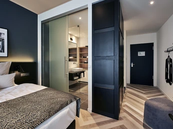 Elegantes Hotelzimmer mit Doppelbett, modernem Interieur, Schiebetür zum Bad und schlichter Dekoration.