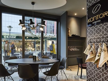 Moderner Cafe-Innenraum mit rundem Tisch, schwarzen Stühlen und Zeitungen an stilvoller Wandhalterung.