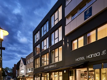 Ein modernes Stadthotel mit beleuchteter Fassade, umgeben von einer Fußgängerzone bei Abenddämmerung.