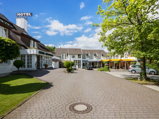 Hotelgebäude mit gepflastertem Vorplatz, dazwischen grüner Rasen und Bäume bei strahlend blauem Himmel.