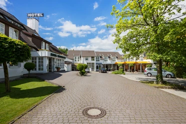 Herford-Hotel Waldesrand.jpg Hotelgebäude mit gepflastertem Vorplatz, dazwischen grüner Rasen und Bäume bei strahlend blauem Himmel.