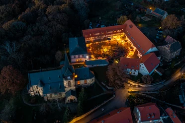 Das Bild zeigt eine Luftaufnahme eines beleuchteten, historischen Gebäudekomplexes in herbstlicher Umgebung, in dem eine Veranstaltung mit zahlreichen Besuchern im Innenhof stattfindet.