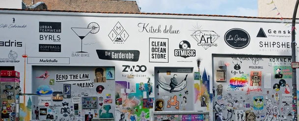 Körnerfeld The picture shows a section of a creatively designed wall in an urban environment. The wall is covered with a variety of colorful posters, signs and graffiti. Some of the logos and signs advertise local businesses.