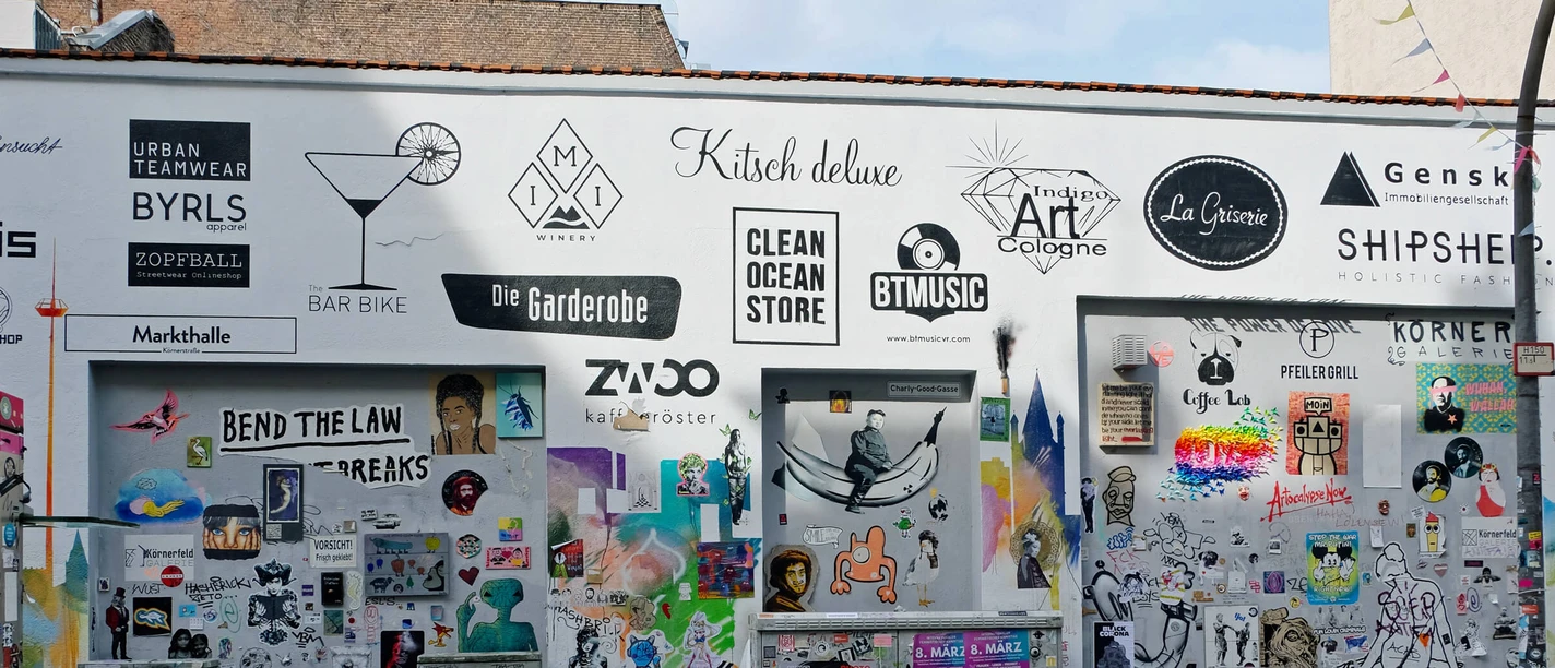 Körnerfeld Auf dem Bild ist ein Abschnitt einer kreativ gestalteten Wand in urbanem Umfeld zu sehen. Die Wand ist mit einer Vielzahl von bunten Postern, Schildern und Graffiti bedeckt. Einige der Logos und Schilder bewerben lokale Geschäfte.