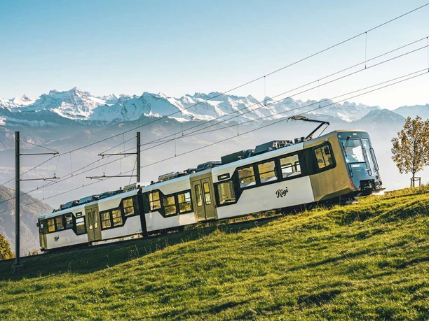 Rigi Bahnen Rigi-Bahn fährt über grüne Bergwiesen vor schneebedeckter Alpenkulisse.