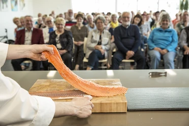 Fischkochstudio Koch präsentiert frisches Lachsfilet vor interessiertem Publikum in moderner Showküche.